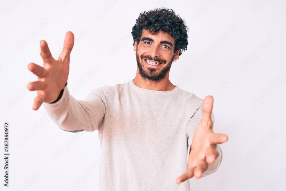 Handsome young man with curly hair and bear wearing casual winter sweater looking at the camera smiling with open arms for hug. cheerful expression embracing happiness. Handsome young man with curly hair and bear wearing casual winter sweater looking at the camera smiling with open arms for hug. cheerful expression embracing happiness.