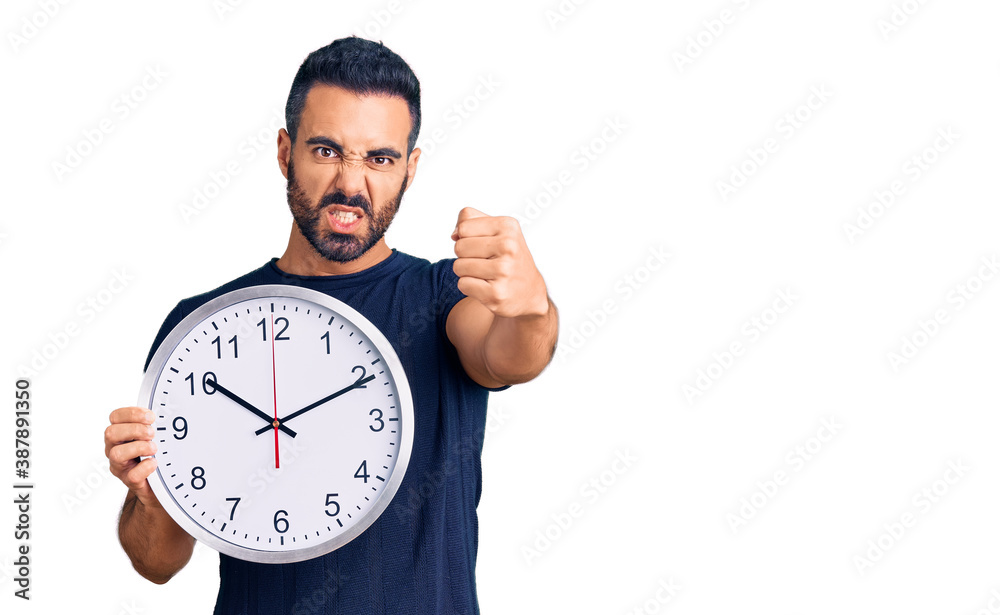Young hispanic man holding big clock annoyed and frustrated shouting with anger, yelling crazy with anger and hand raised Young hispanic man holding big clock annoyed and frustrated shouting with anger, yelling crazy with anger and hand raised