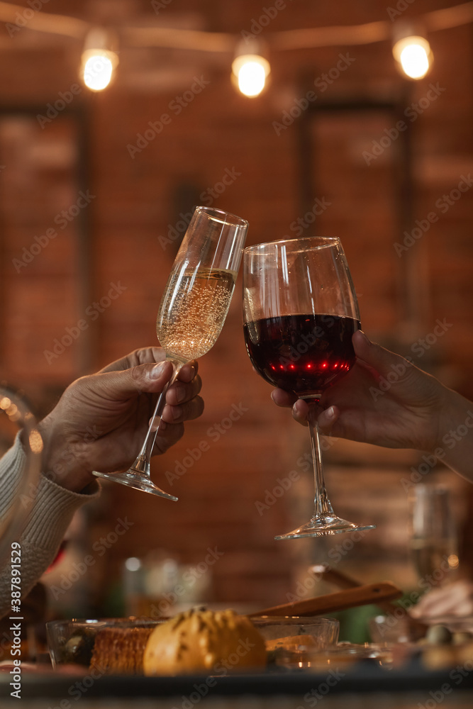Vertical close up of unrecognizable loving couple clinking glasses while enjoying dinner date in romantic ambience