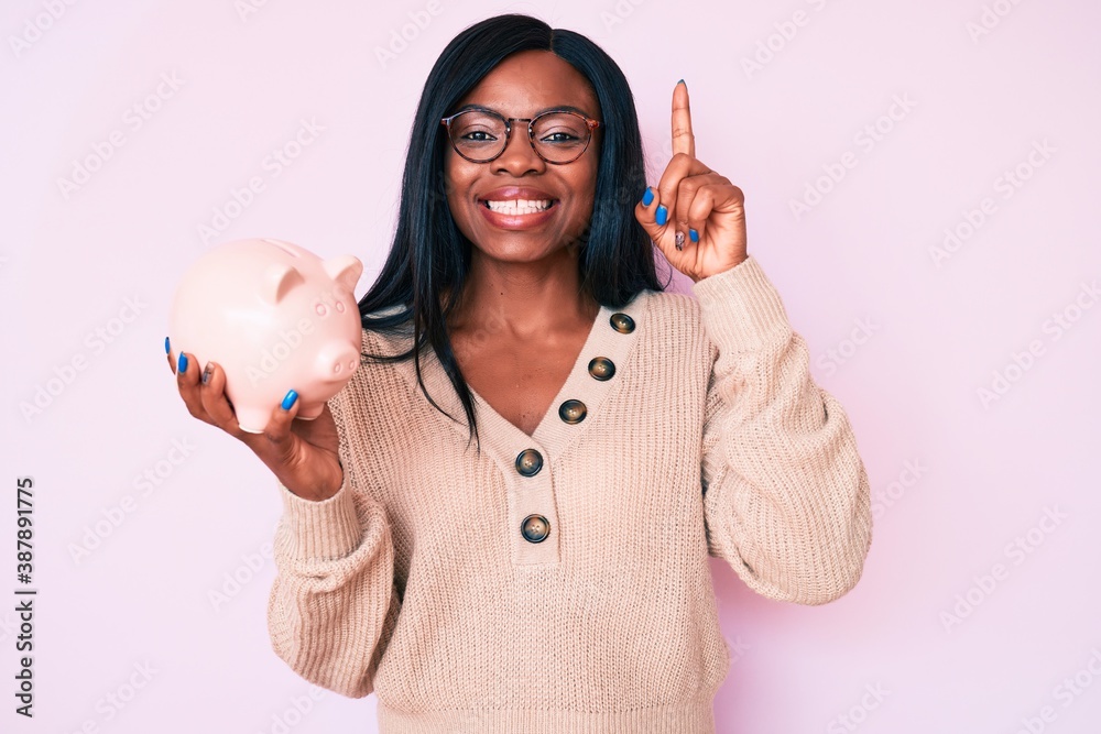 Young african american woman holding piggy bank smiling with an idea or question pointing finger with happy face, number one Young african american woman holding piggy bank smiling with an idea or question pointing finger with happy face, number one