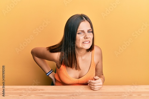 Young caucasian woman wearing casual clothes sitting on the table suffering of backache, touching back with hand, muscular pain