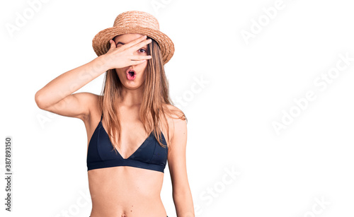 Young beautiful girl wearing bikini and hat peeking in shock covering face and eyes with hand, looking through fingers with embarrassed expression.