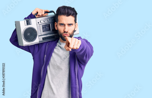 Young handsome man with beard listening to music using vintage boombox pointing with finger to the camera and to you, confident gesture looking serious