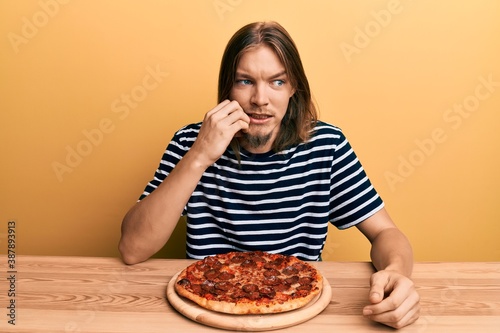 Handsome caucasian man with long hair eating tasty pepperoni pizza looking stressed and nervous with hands on mouth biting nails. anxiety problem.