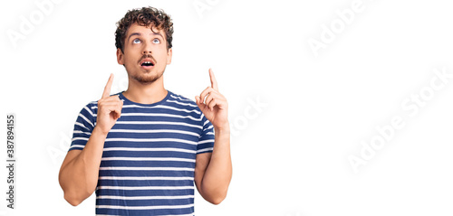 Young handsome man with curly hair wearing casual clothes amazed and surprised looking up and pointing with fingers and raised arms.