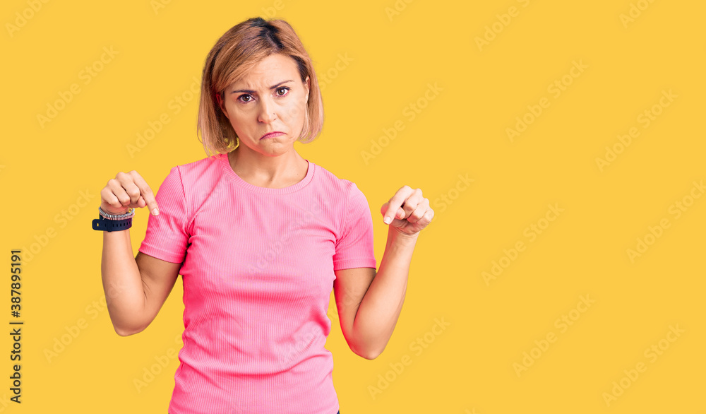 Young blonde woman wearing sportswear pointing down looking sad and upset, indicating direction with fingers, unhappy and depressed.