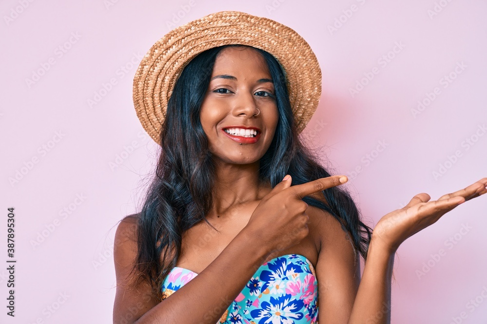 Young indian girl wearing bikini and summer hat amazed and smiling to the camera while presenting with hand and pointing with finger. Young indian girl wearing bikini and summer hat amazed and smiling to the camera while presenting with hand and pointing with finger.