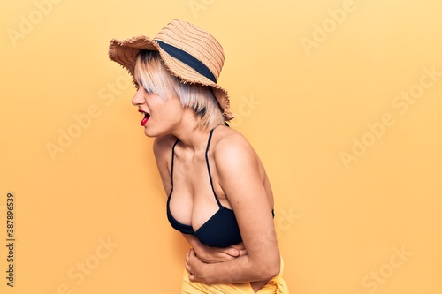 Young beautiful blonde woman wearing bikini and summer hat with hand on stomach because nausea, painful disease feeling unwell. ache concept. photo