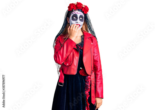 Woman wearing day of the dead costume over background looking stressed and nervous with hands on mouth biting nails. anxiety problem.