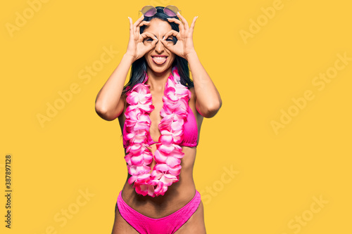 Beautiful hispanic woman wearing bikini and hawaiian lei doing ok gesture like binoculars sticking tongue out, eyes looking through fingers. crazy expression.