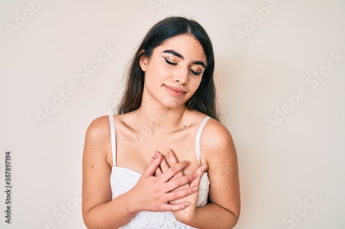 Brunette teenager girl posing elegant smiling with hands on chest with closed eyes and grateful gesture on face. health concept.