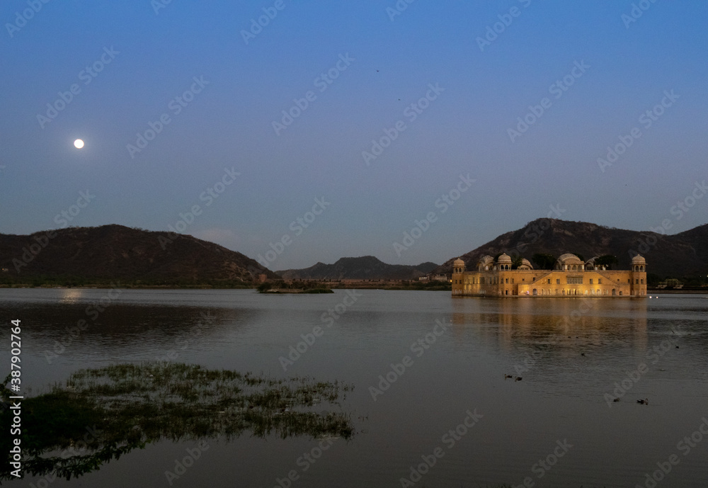 night shot of the moon rising and a floodlit jal mahal palace in jaipur