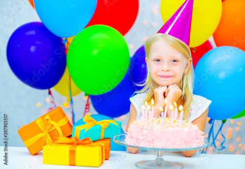 Little girl in party cap looks happily at cake with birthday candles