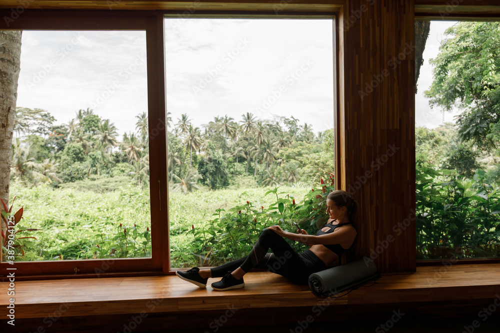 Young woman in sport clothes sit near the window with tropical view otdoor and palm trees, using mobile phone. Rolled yoga mat lie ear her, waiting for trainining Young woman in sport clothes sit near the window with tropical view otdoor and palm trees, using mobile phone. Rolled yoga mat lie ear her, waiting for trainining