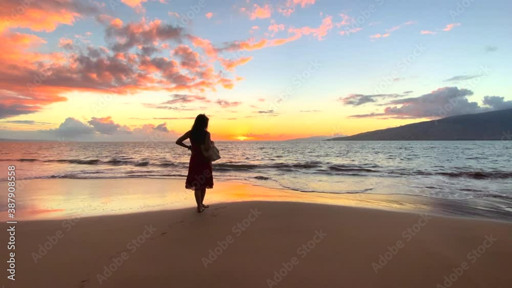 Silhouette of woman in a dress standing on a sandy tropical beach at sunset in a medium shot with gentle waves rolling in and part of an island in the distance and orange color on the ocean Silhouette of woman in a dress standing on a sandy tropical beach at sunset in a medium shot with gentle waves rolling in and part of an island in the distance and orange color on the ocean