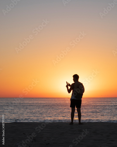 Man silhouette in a beautiful beach while taking a photo with his cellphone