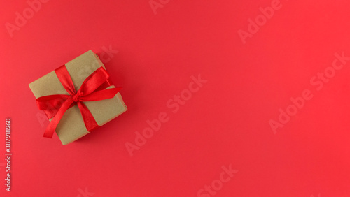 Gift box wrapped in a craft paper with red ribbon and bow on red background. Monochrome festive flat lay with copy space.