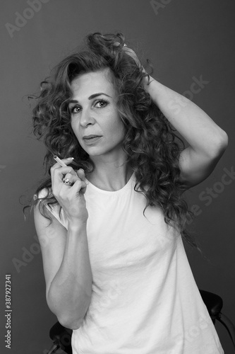 A beautiful brunette girl with long curly hair in a white T-shirt smokes in the studio on a gray background  bw