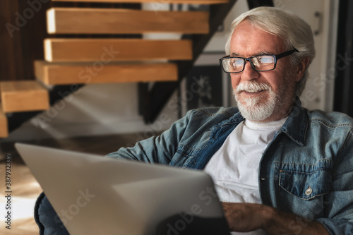 Elderly man with glasses with gray hair and a beard sits at home on the sofa and works or browses social networks on  laptop