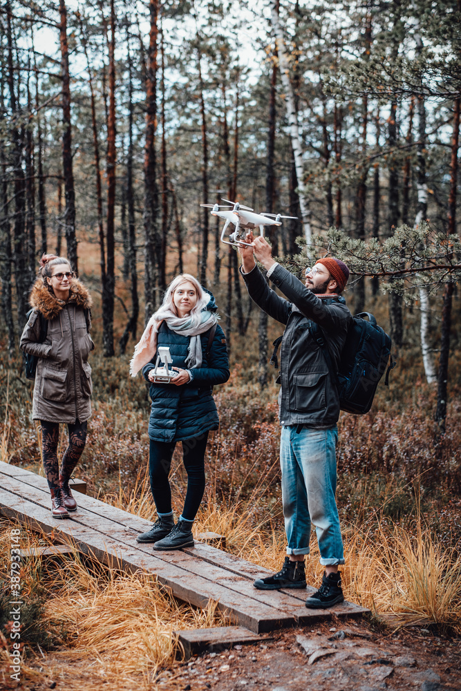 Cheerful group of students a guy he holds quadcopter with raised hands and two girls they stay near him in lovely autumn forest.