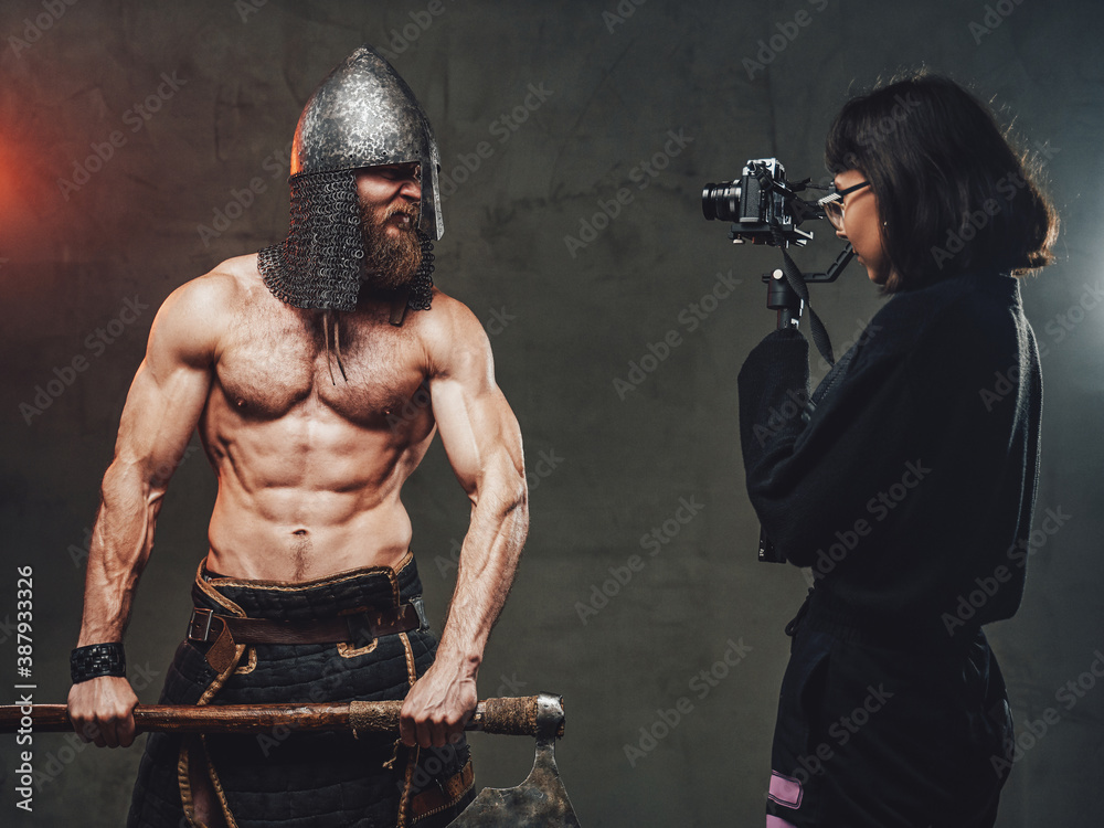 Shirtless and muscular guy with axe and helmet in fashion of savage viking poses for camera while photographer shots him.