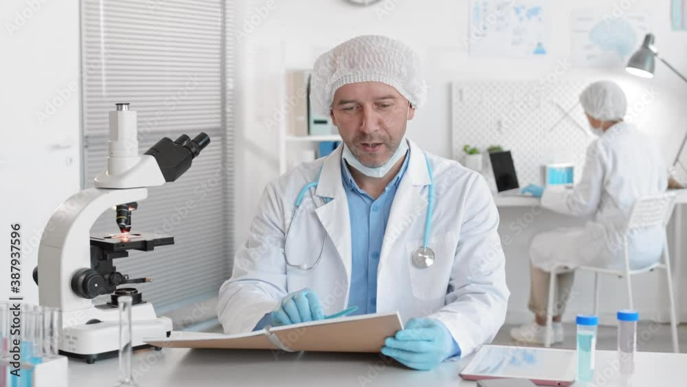 Medium POV of blue-eyed adult Cacasian male doctor sitting by desk in busy laboratory wearing protection, taking face mask off and talking to person by video call Medium POV of blue-eyed adult Cacasian male doctor sitting by desk in busy laboratory wearing protection, taking face mask off and talking to person by video call