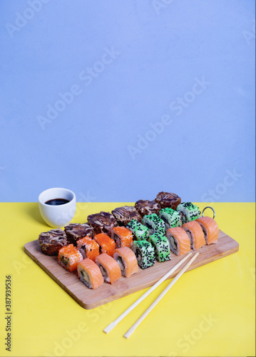 Sushi roll with soy sauce and chopsticks on a wooden stand on a bright yellow-blue background. Sushi menu.