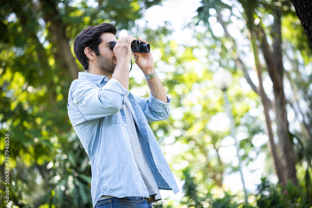 Young handsome man looking natural and using binoculars in public park with a happy face standing and smiling. Concept of nature and wildlife studies.