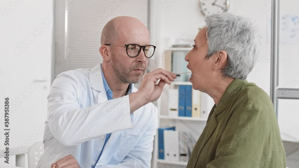 Medium slow motion of Caucasian male therapist inserting cotton swab into mouth of senior female patient for virus testing in bright hospital office