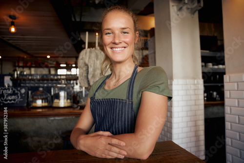 Cheerful young female waitress wearing apron standing leaning on table in cafe