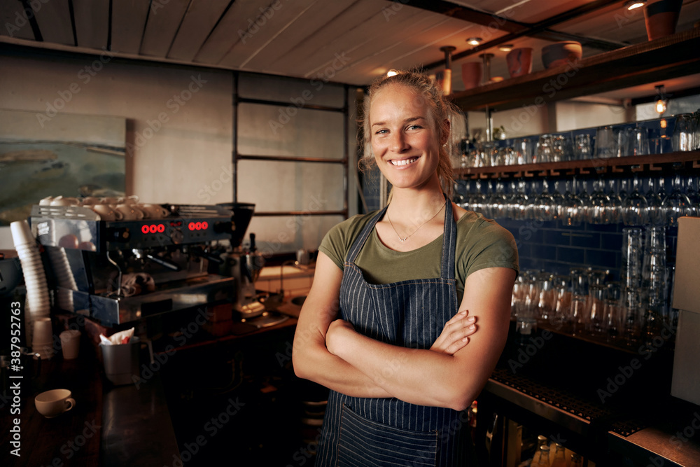 Portrait of young female waitress wearing apron standing behind counter in cafe with crossed hands