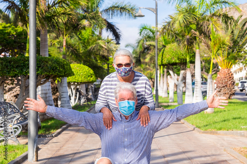 Couple of two senior grandparents having fun together in outdoor park wearing surgical mask due to coronavirus pandemic. Concept of funny elderly people and new normal life with virus Covid-19