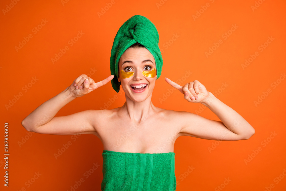 Photo portrait of woman pointing fingers at golden eye patches wearing green bath towel isolated on vivid orange colored background Photo portrait of woman pointing fingers at golden eye patches wearing green bath towel isolated on vivid orange colored background