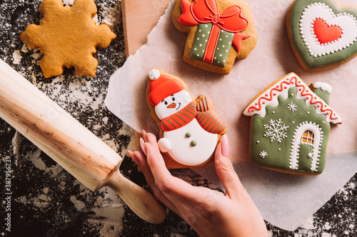 Christmas bakery. Festive decorated pastry. Homemade biscuits. Unrecognizable female hand holding gingerbread icing snowman figured cookies on oilpaper flour powder rolling pin on desk. photo