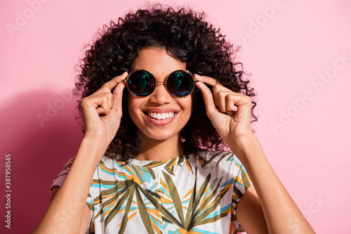 Portrait photo of black skinned curly woman wearing round stylish sunglass smiling isolated on pastel pink color background © deagreez