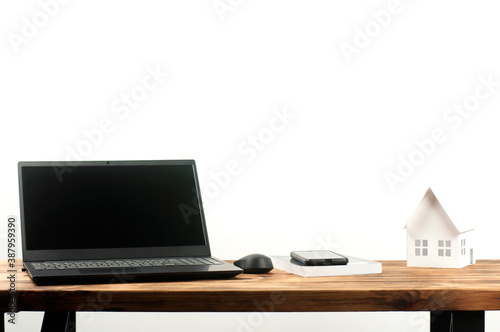Laptop and computer mouse. Phone and book. Miniature paper house. On a wooden table.
