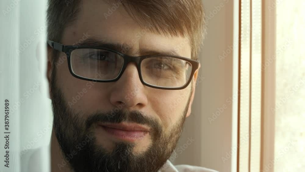 Close up of young bearded man with glasses by window drinks cup of fresh hot coffee, looks at camera, smiles and laughs. Morning coffee concept. Close up of young bearded man with glasses by window drinks cup of fresh hot coffee, looks at camera, smiles and laughs. Morning coffee concept.
