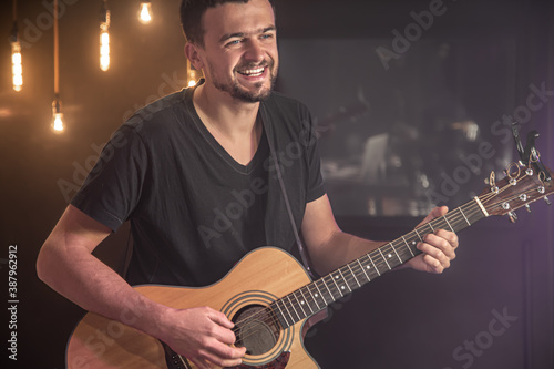 Happy guitarist in a black t-shirt plays an acoustic guitar at a concert against a blurred black background.