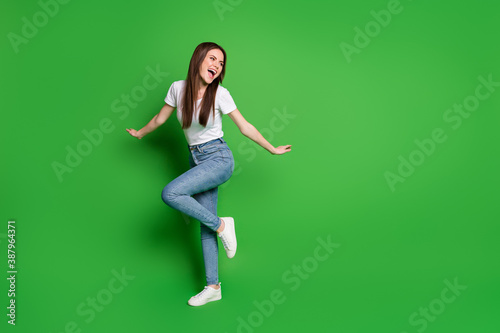 Full length photo of adorable dreamy young lady dancing wear jeans t-shirt footwear isolated on green background