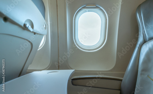 Plane window with white sunlight. Empty plastic airplane tray table at seat back. Economy class airplane window. Inside of commercial airline. Seat with armchair. Leather seat of economy class plane. © Artinun