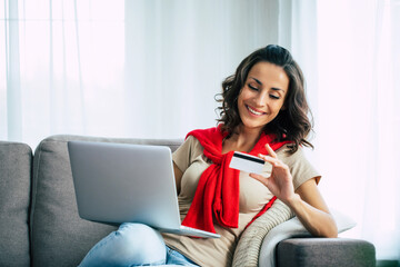 Beautiful happy young woman in casual clothes is sitting on the couch at home and doing online shopping on black friday.