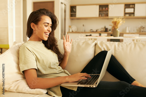 Cute student girl learning distantly from home, sitting on sofa with portable computer on lap, making greeting gesture, waving hand, saying Hello there. Communication and electronic gadgets concept