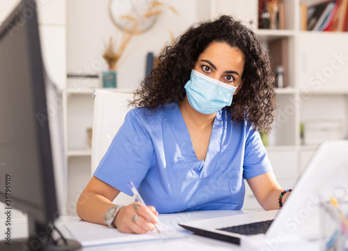 Portrait of young female doctor in face mask working on laptop at office in hospital
