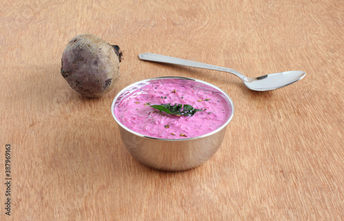 Beetroot raita or pachadi, a healthy Indian vegetarian side dish for food like chapati, roti, naan, and paratha, is made from ingredients like beetroot gratings and curd, in a steel bowl on a wooden b