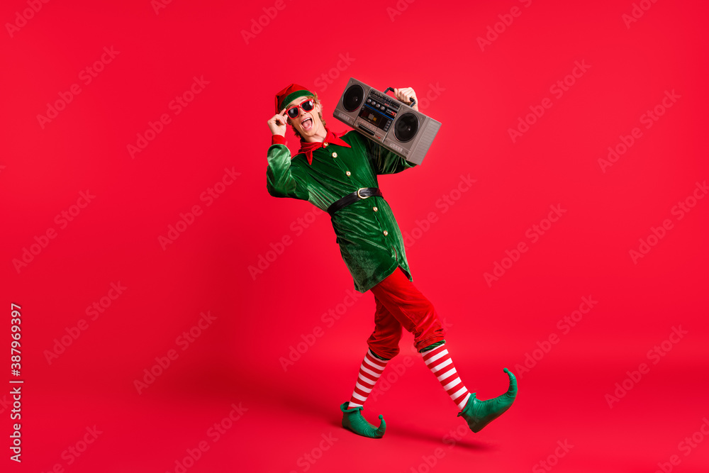 Full size profile side photo of crazy elf hold boom box go wear green costume isolated over red shine color background Full size profile side photo of crazy elf hold boom box go wear green costume isolated over red shine color background