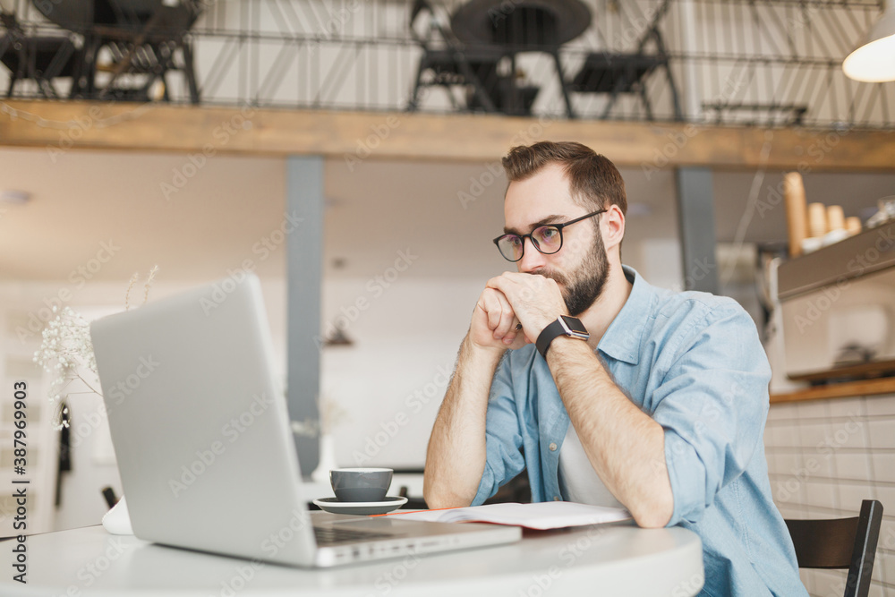 Young man in eyeglasses sitting alone at table in coffee shop cafe working or studying on laptop pc computer, relaxing in restaurant during free time indoors. Freelance mobile office business concept. Young man in eyeglasses sitting alone at table in coffee shop cafe working or studying on laptop pc computer, relaxing in restaurant during free time indoors. Freelance mobile office business concept.