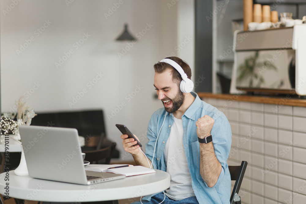 Happy young man sit at table in coffee shop cafe restaurant indoors working studying on laptop listening music with headphones cell phone doing winner gesture Freelance mobile office business concept.