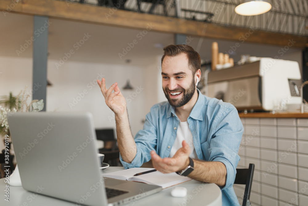 Fototapeta premium Cheerful young man in air pods sitting alone at table in coffee shop cafe restaurant indoors working or studying on laptop pc computer making video call. Freelance mobile office business concept.