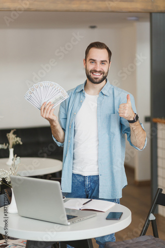 Smiling man stand near table in coffee shop cafe restaurant indoors working studying on laptop hold fan of cash money in dollar banknotes showing thumb up. Freelance mobile office business concept.