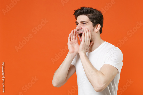 Crazy displeased young man wearing basic casual blank empty white t-shirt standing screaming with hands gesture near mouth looking aside isolated on bright orange colour background, studio portrait.